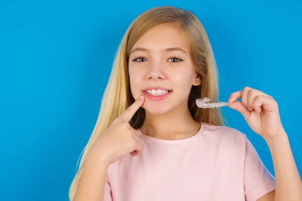 Caucasian kid girl wearing pink shirt against blue wall holding an invisible aligner and pointing to her perfect straight teeth. Dental healthcare and confidence concept.