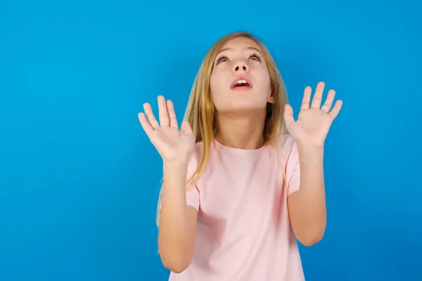 白种人小女孩穿着粉色衬衫靠着蓝色的墙 手心前倾 恐惧地看着天花板 试图保护自己不受看不见的危险的伤害 张开嘴 — 图库照片