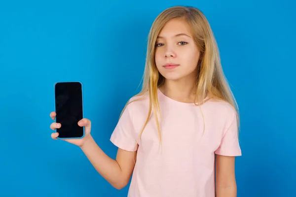 白人の子供女の子身に着けていますピンクシャツに対して青壁ホールド新しい携帯電話と神秘的な脇ショーブロンド表示の現代の携帯電話 — ストック写真