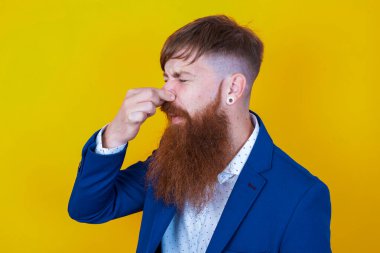 Sakallı, beyaz, mavi ceketli adam sarı arka planda poz veriyor.