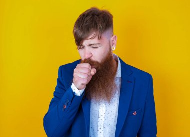 Sakallı, beyaz, mavi ceketli adam sarı arka planda poz veriyor.