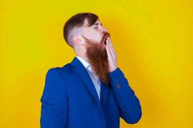 Sakallı, beyaz, mavi ceketli adam sarı arka planda poz veriyor.