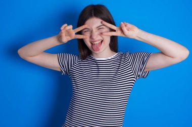 Neşeli, pozitif, genç bir kadın gözlerinin yanında V işareti gösteriyor.  