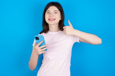 Portrait of young brunette woman using and texting with smartphone  happy with big smile doing ok sign, thumb up with fingers, excellent sign