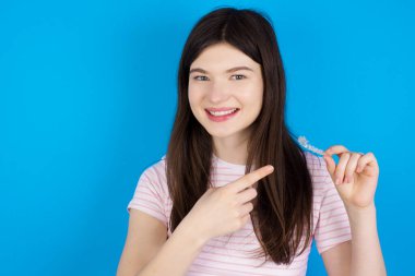 young brunette woman holding an invisible aligner and pointing at it. Dental healthcare and confidence concept.