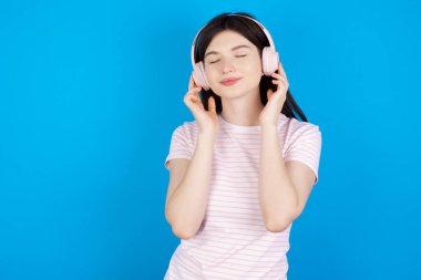 Pleased young brunette woman enjoys listening pleasant melody keeps hands on stereo headphones closes eyes. Spending free time with music