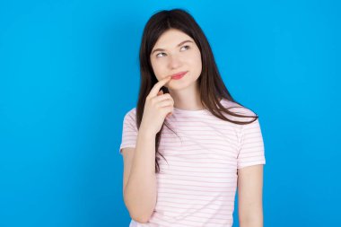 Lovely dreamy young brunette woman keeps finger near lips looks aside copy space.