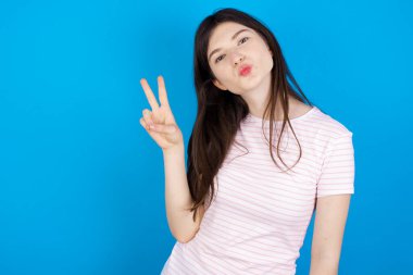 young brunette woman makes peace gesture keeps lips folded shows v sign. Body language concept