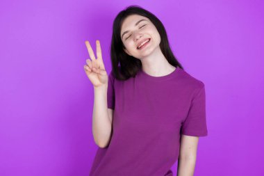 Genç esmer kadın, gülümseyen mutlu bir yüzle kameraya göz kırpıyor zafer işareti yapıyor. İki numara..