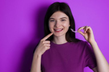 young brunette woman holding an invisible aligner and pointing to her perfect straight teeth. Dental healthcare and confidence concept.