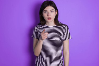 Shocked young brunette woman points at you with stunned expression