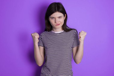 Irritated young brunette woman blows cheeks with anger and raises clenched fists expresses rage and aggressive emotions. Furious model