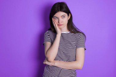 Sad lonely young brunette woman touches cheek with hand bites lower lip and gazes with displeasure. Bad emotions