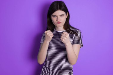 Displeased annoyed young brunette woman clenches fists, gestures pissed, ready to revenge, looks with aggression at camera stands full of hate, being pressured