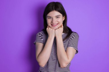 Satisfied young brunette woman touches chin with both hands, smiles pleasantly, rejoices good day with lover