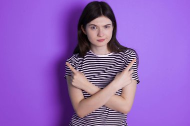 Serious young brunette woman crosses hands and points at different sides hesitates between two items. Hard decision concept