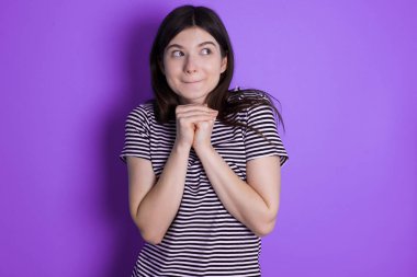 Curious young brunette woman keeps hands under chin bites lips and looks with interest aside.