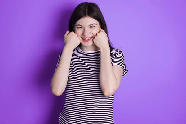 Happy young brunette woman keeps fists on cheeks smiles broadly and has positive expression being in good mood