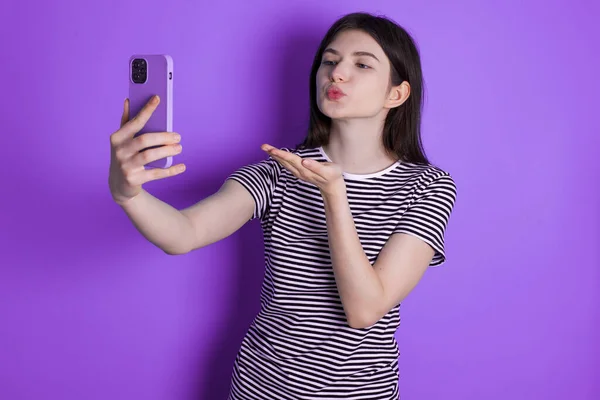 若いブルネットの女性はスマートフォンのカメラで空気のキスを吹くと自画撮りを取ります オンライン通話を介してMwahを送信 — ストック写真