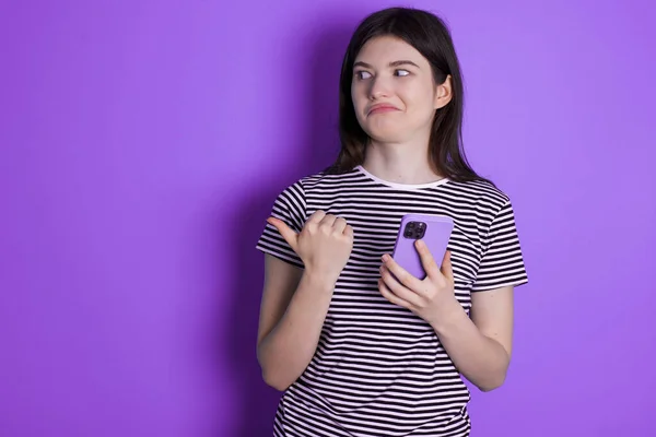 young brunette woman points thumb away and shows blank space aside, holds mobile phone for sending text messages.