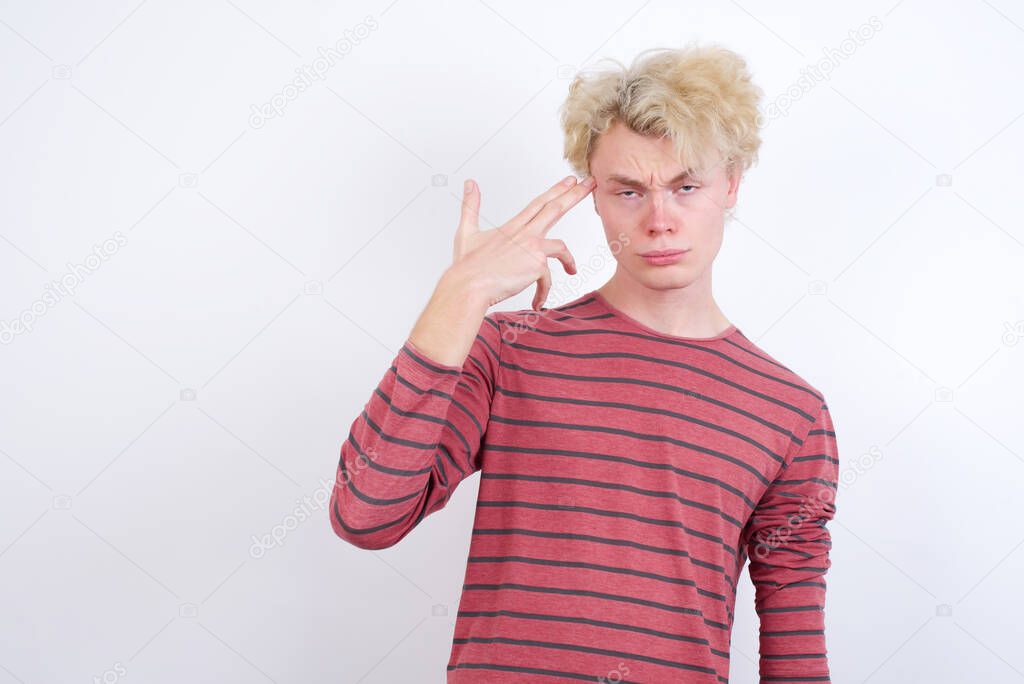 Unhappy young man makes suicide gesture and imitates gun with hand, curves lips, keeps two fingers on temple