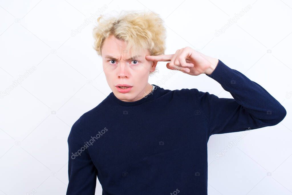 Unhappy young caucasian man makes suicide gesture and imitates gun with hand, curves lips keeps two fingers on temple, shoots, being tired of everything,