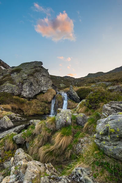 Suddigt flödande vattenfall i Alperna med färgglada himmel och moln vid skymningen, bland klippor, stenblock och gräs. Liten bäck i idyllisk oförorenad miljö, lång exponering. — Stockfoto