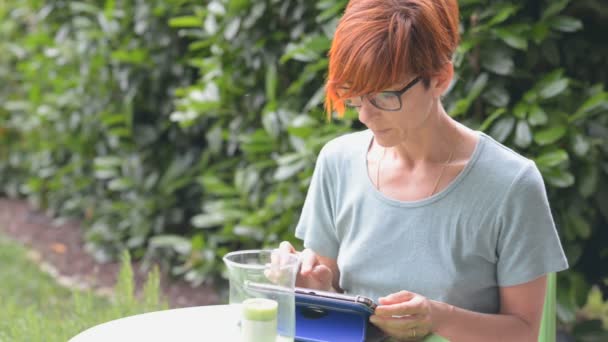 Kadın gözlük ve gündelik Garnitür Telleri açık havada çiçekli bahçesinde otururken tablet ile çalışma. Doğal gün ışığı alan, gerçek insanlar. Her yerde çalışma kavramı yeni bağlantı teknolojisi ile. — Stok video