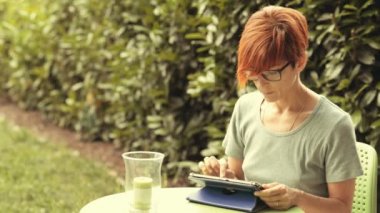 Açık havada çiçekli bahçesinde otururken tablet ile çalışma gözlüklü kadın. Tonlu görüntü, retro vintage tarzı, gerçek insanlar. Her yerde çalışma kavramı yeni bağlantı teknolojisi ile.