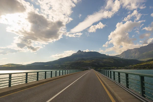 Dlážděná dvoucestná silnice na mostě přes jezero v malebné krajině a na náladové obloze. Panoramatický výhled z kamery na auto. Letní dobrodružství a výlet na dovolenou. — Stock fotografie