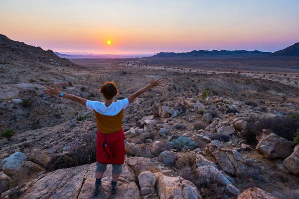 Turista observando la impresionante vista del valle estéril en el desierto de Namib, majestuosa atracción de visitantes en Namibia, África. Naranja rojo violeta claro cielo en el horizonte, brillantes rocas y cañón en el para — Foto de Stock