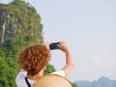 Vietnam Halong Körfezi 'nde telefon kullanan geleneksel şapkalı bir kadın. Ha Long Körfezi kaya zirvelerinde seyahat eden bir turist. Beyaz kadın ünlü şehir simgesinde tatilde eğleniyor..