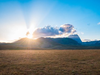 Kayalık dağlar, dağlık araziler ve otlaklar üzerinde gün batımı manzarası. Campo Imperatore, Gran Sasso, Apennines, İtalya. Gökyüzündeki güneş ışınları ve renkli bulutlar dramatik dağ sırtlarında.