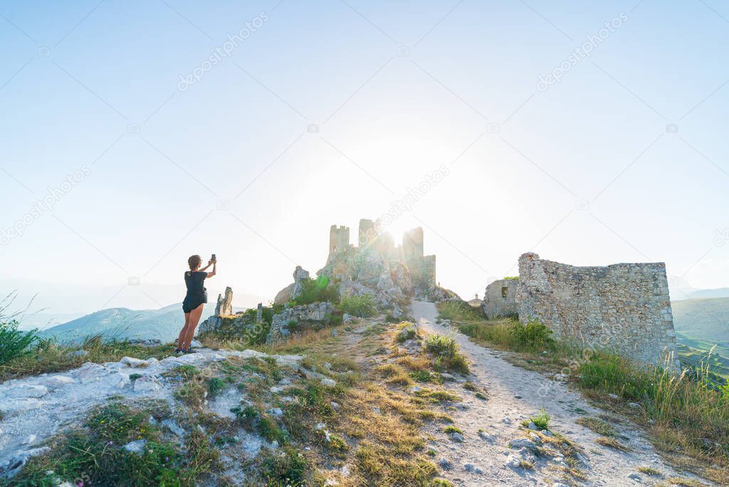 Woman selfie at castle ruins on mountain top at Rocca Calascio, italian travel destination, landmark in the Gran Sasso National Park, Abruzzo, Italy. Clear blue sky sun burst in backlight