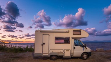 Gün batımında deniz kenarında, dramatik bulutlarla dolu renkli bir gökyüzünün altında park edilmiş bir karavan. Huzurlu bir akşam atmosferi, seyahat, macera, özgürlük ve açık hava yaşam temaları için mükemmel.