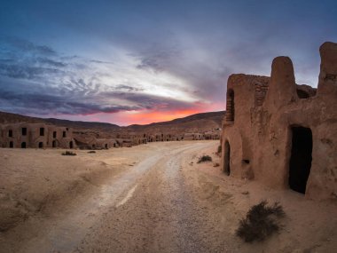 Güney Tunus 'ta gün batımında Ksar Hallouf' un geniş açılı görüntüsü tozlu bir yol, dramatik bulutlar ve sıcak çöl ışığı boyunca antik çamur tuğlalı evler gösteriyor.