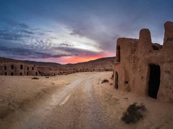 Güney Tunus 'ta gün batımında Ksar Hallouf' un geniş açılı görüntüsü tozlu bir yol, dramatik bulutlar ve sıcak çöl ışığı boyunca antik çamur tuğlalı evler gösteriyor.