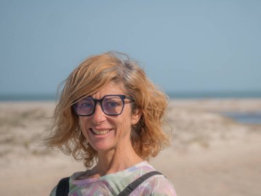 Gözlüklü, gülümseyen olgun bir kadın açık deniz manzarasında ayakta duruyor. Özgüven, özgünlük ve aktif yaşam tarzını aktaran doğal portre, refah, seyahat ve olgun yaşam tarzı kavramları için idealdir.