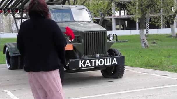 Pembe etekli ve siyah ceketli orta yaşlı bir kadın, rüzgarlı bir havada savaş zamanı Katyusha arabasının anıtına çiçek bırakıyor. — Stok video