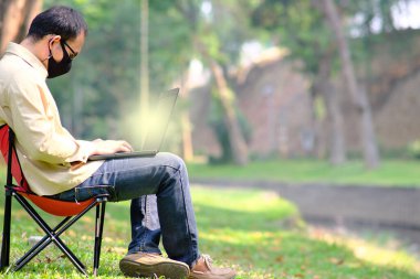 Genç adam bankta oturuyor ve parkta dizüstü bilgisayarla çalışıyor.