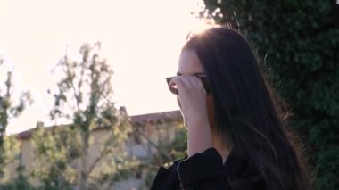 Genç Yetişkin, güneş gözlüğü kaldırıyor rüzgarda uçuşan saçlar. Güneş ışınlarında ve alevlerde kız mutlu bir şekilde gülümsüyor. Slow Motion Shot 180fps