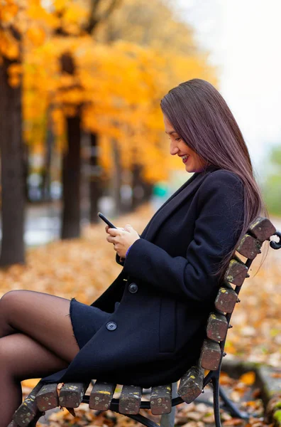 秋の若い女性の肖像画 暖かい光と落ち葉の風景 彼女はスマートフォンを使って微笑んだ — ストック写真