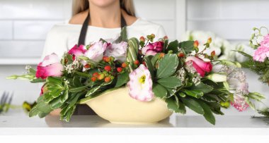 Çiçekçi elinde güller, ranunculus, hiperikum meyveleri ve çeşitli yeşil yapraklar içeren renkli bir çiçek aranjmanı tutuyordu. Çiçekçide beyaz bir mermer tezgahın arkasında duruyordu.