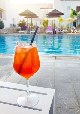 Canlı turuncu Aperol Spritz kokteyli, buzlu ve pipetli, beyaz bir masada, güneşli yaz güneşinin altında dinlendirici mavi bir yüzme havuzuna bakıyor.