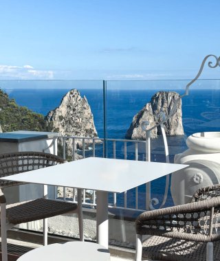 Güneşli bir günde, açık mavi Akdeniz 'e karşı ikonik Faraglioni deniz yığınlarının nefes kesici manzarasını sunan manzaralı terasta modern masa ve sandalyeler.