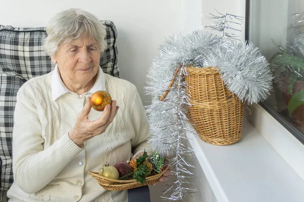 Sandalyede oturan ve elinde Noel oyuncağı tutan gri saçlı son sınıf öğrencisi kadın. Yaşlı kadın Noel ve yeni yıla hazırlanıyor..