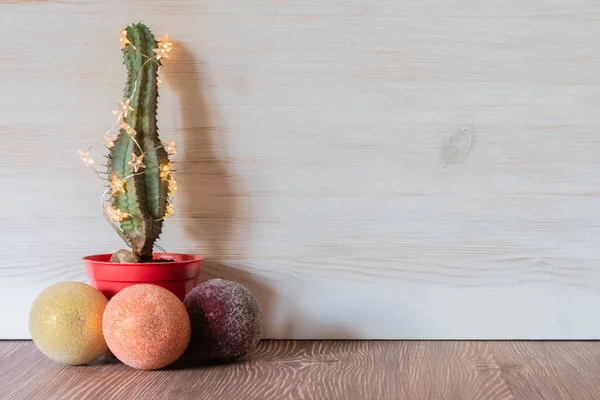 Christmas cactus tree with shining garland and baubles around on wooden background. Cactus as alternative Christmas tree with copy space. Tropical Christmas mood.