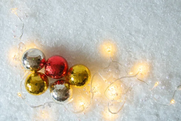 Christmas balls and shining garland on snow imitation background. Christmas and New Year decoration of glowing garlands and baubles on snow. Top view. Copy space.
