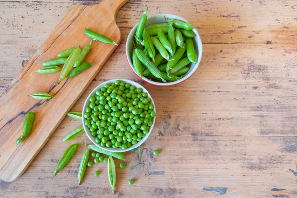 Fresh green peas in bowl and pods placed on wooden board on rustic table copy space. Pods of pea on wood cutting board and bowl of fresh green peas.