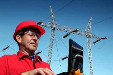 Kırmızı başlıklı elektrik mühendisi yüksek voltajlı elektrik hatlarının önünde dizüstü bilgisayar kullanıyor.